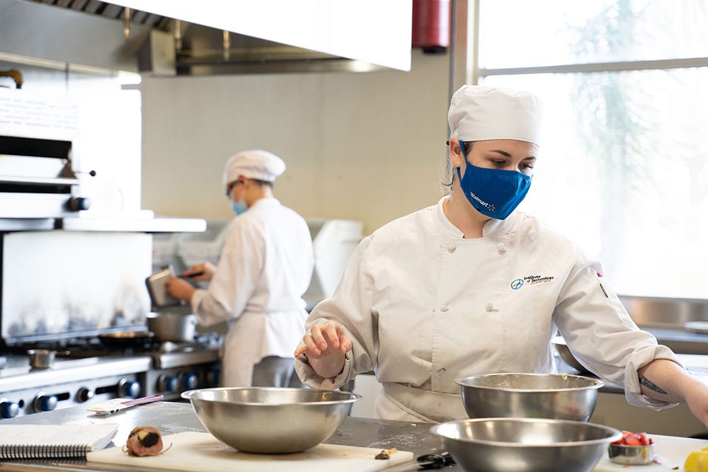 Woman prepping kitchen while another person works over a grill